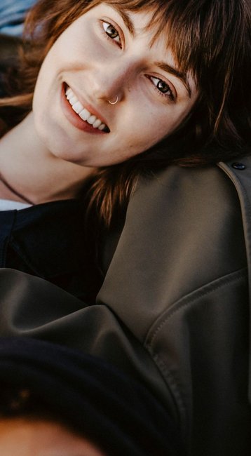 Fünf Personen liegen auf dem Boden, lächelnd und in einer entspannten Position. Sie haben unterschiedliche Haarfarben und tragen lässige Kleidung. Ihre Hände sind miteinander verbunden, was eine freundliche Atmosphäre vermittelt.