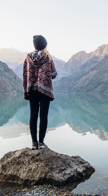 Eine Person steht auf einem Felsen am Ufer eines ruhigen Sees, umgeben von Bergen. Das Wasser spiegelt die Landschaft wider, während die Person in Richtung der Berge blickt.