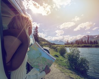Eine Person lehnt aus einem Fahrzeugfenster und hält eine Landkarte, während sie auf einen Fluss und die umliegenden Berge blickt. Die Szene ist von natürlichem Licht und einer klaren, sonnigen Atmosphäre geprägt.