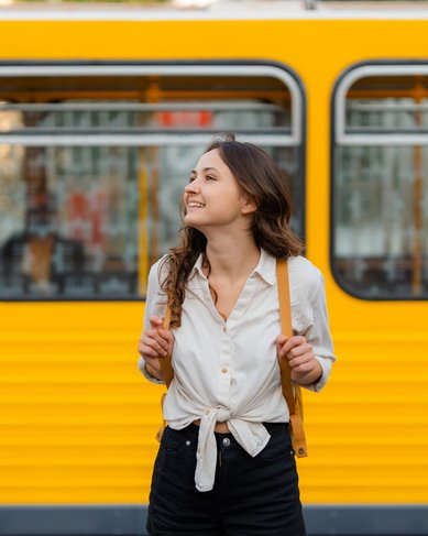 Eine junge Frau mit lockigem Haar trägt ein weißes Hemd und schwarze Shorts. Sie steht vor einem gelben Zug und lächelt, während sie einen Rucksack mit beiden Händen hält.