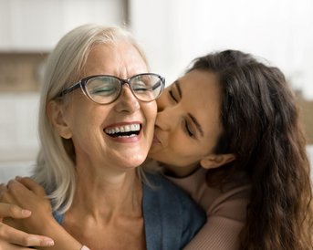 Eine ältere Frau mit Brille lächelt, während eine jüngere Frau sie von hinten umarmt und ihr einen Kuss auf die Wange gibt.